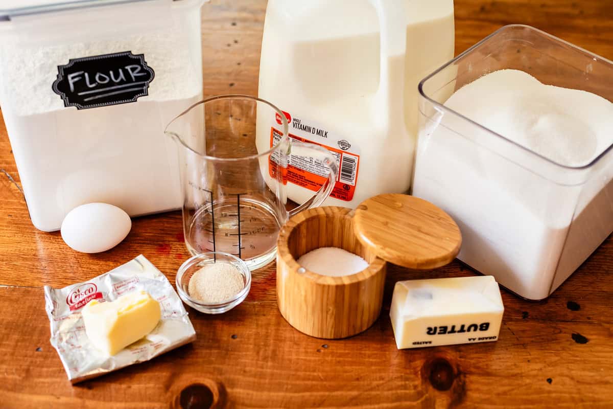 Baking ingredients on a wooden table: flour, milk, sugar, salt, butter, an egg, yeast, and a measuring cup with water. Each item is in its own container or packaging, ready for use.