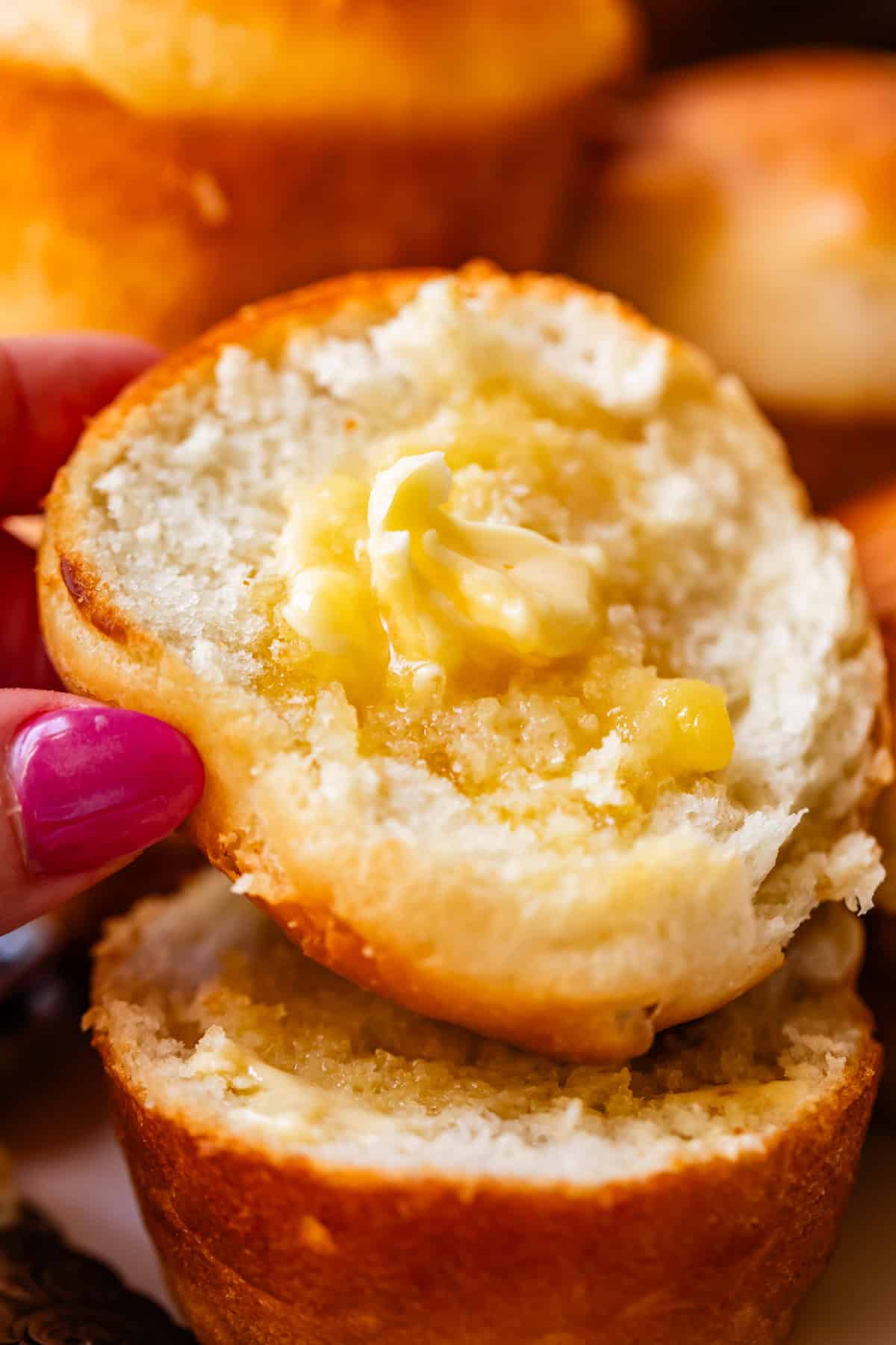 A hand with pink nail polish holds a warm, split muffin topped with melting butter. More muffins are visible in the background.