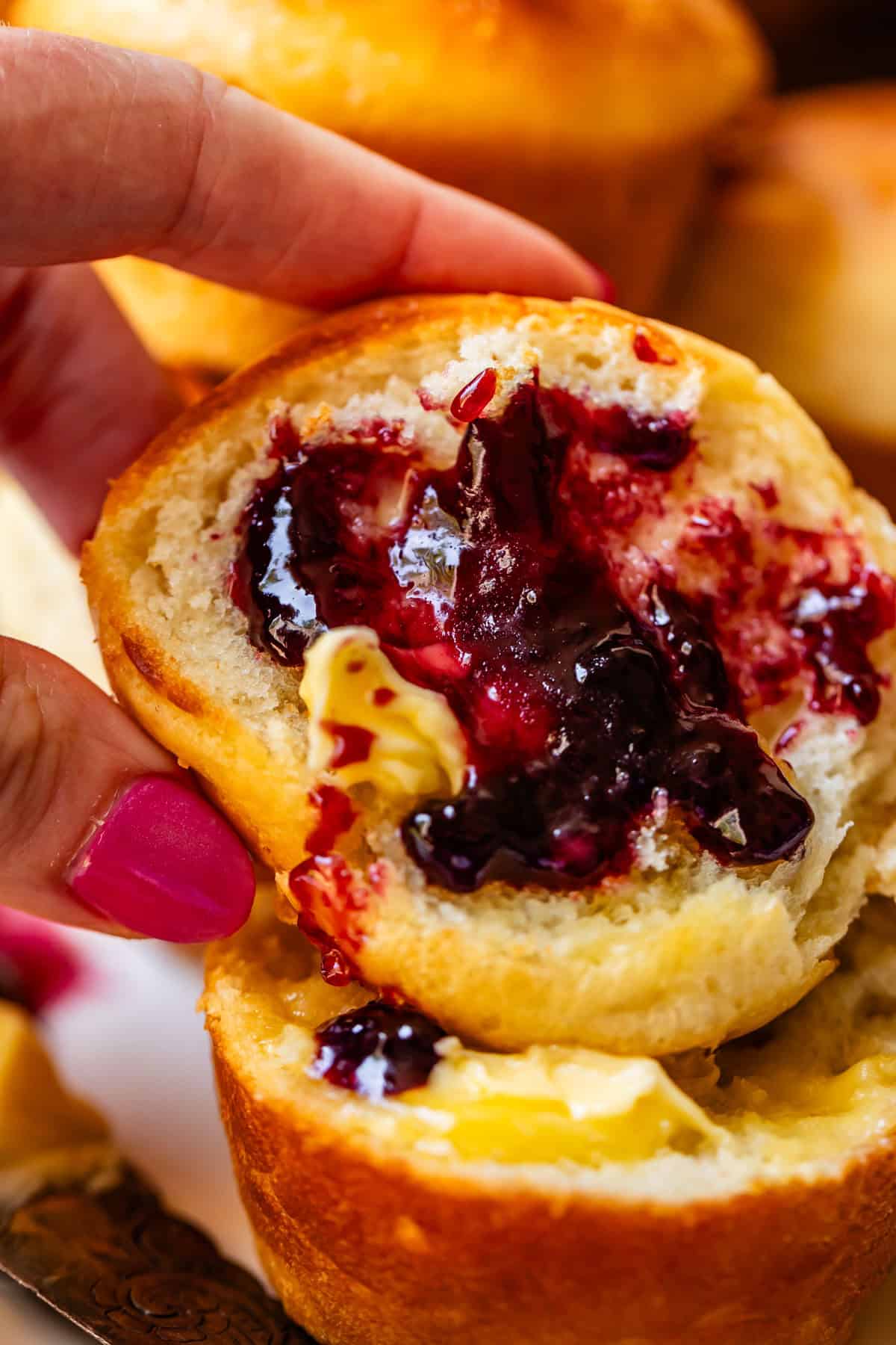 A close-up of a hand holding a muffin split open, spread with butter and dark berry jam. The persons fingernails are painted pink. The muffin looks fluffy and golden brown.
