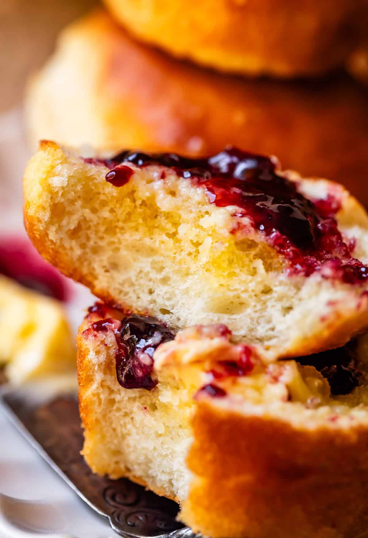A close-up of a fluffy biscuit split open, topped with melting butter and glossy, dark red berry jam, with more biscuits blurred in the background.