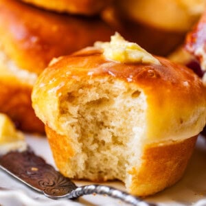 A golden brown dinner roll with a bite taken out sits on a white plate, topped with a pat of melting butter. More rolls and a butter knife are visible in the background.