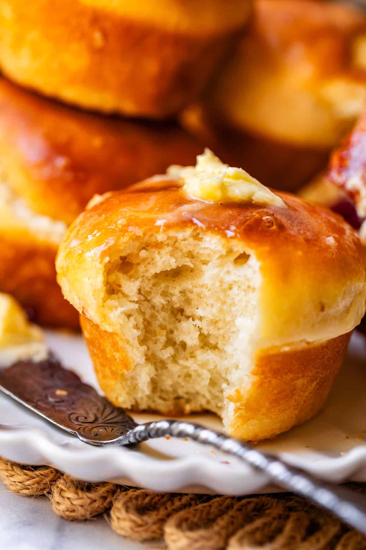 A golden brown dinner roll with a bite taken out sits on a white plate, topped with a pat of melting butter. More rolls and a butter knife are visible in the background.