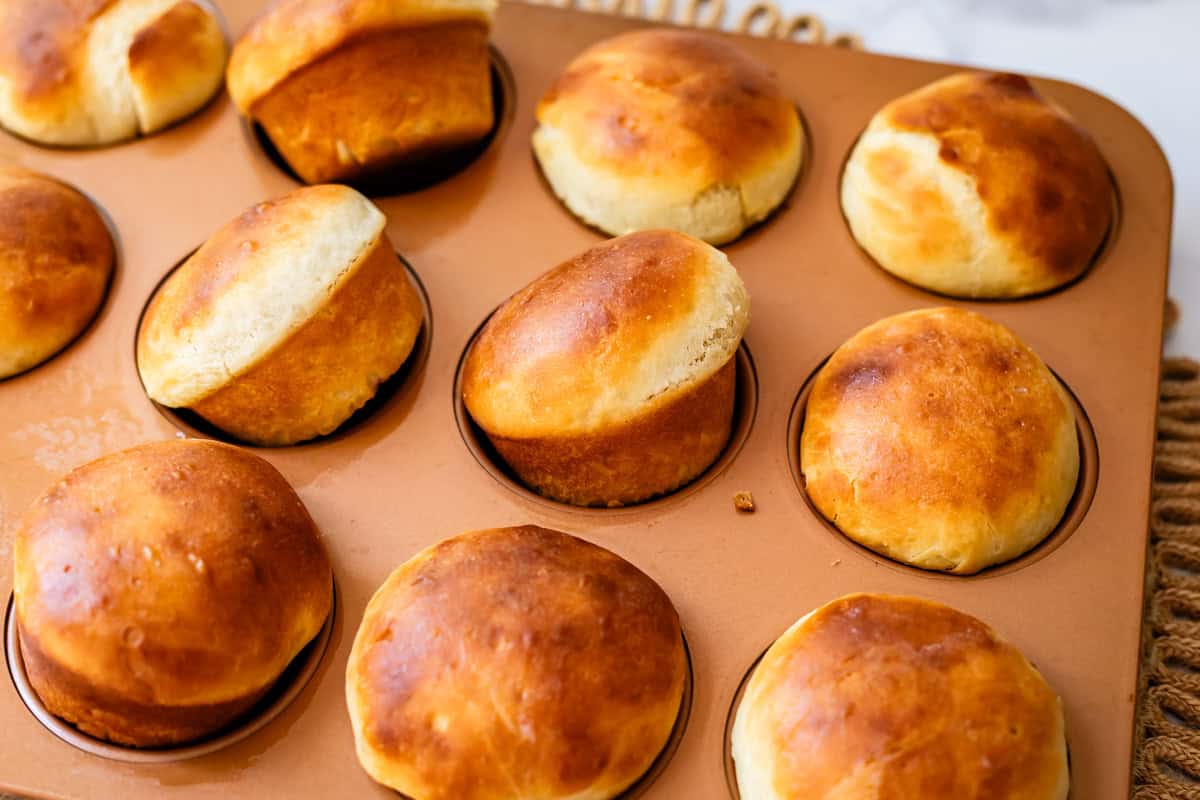 Golden brown rolls fresh out of the oven are shown in a muffin tin, with some rolls puffed up more than others. The rolls have a shiny, slightly uneven surface and a soft, fluffy appearance.