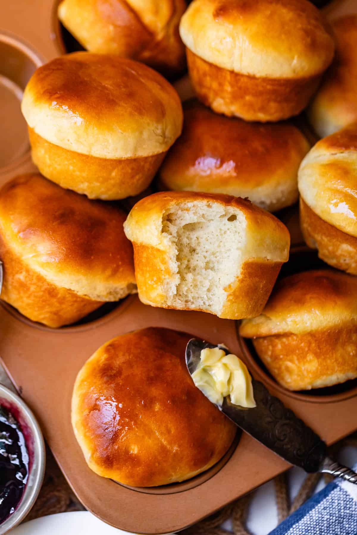 A batch of shiny, golden brown rolls in a muffin tin, with one roll pulled apart to show its soft interior. A knife with butter spreads on a roll, and a jar of jam is partially visible nearby.