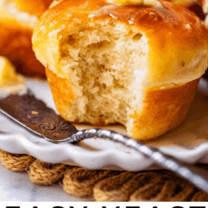 A close-up of a golden brown dinner roll with a soft, fluffy interior, partially eaten and topped with melted butter, resting on a plate with a butter knife. Text below reads, Easy Yeast Dinner Rolls.