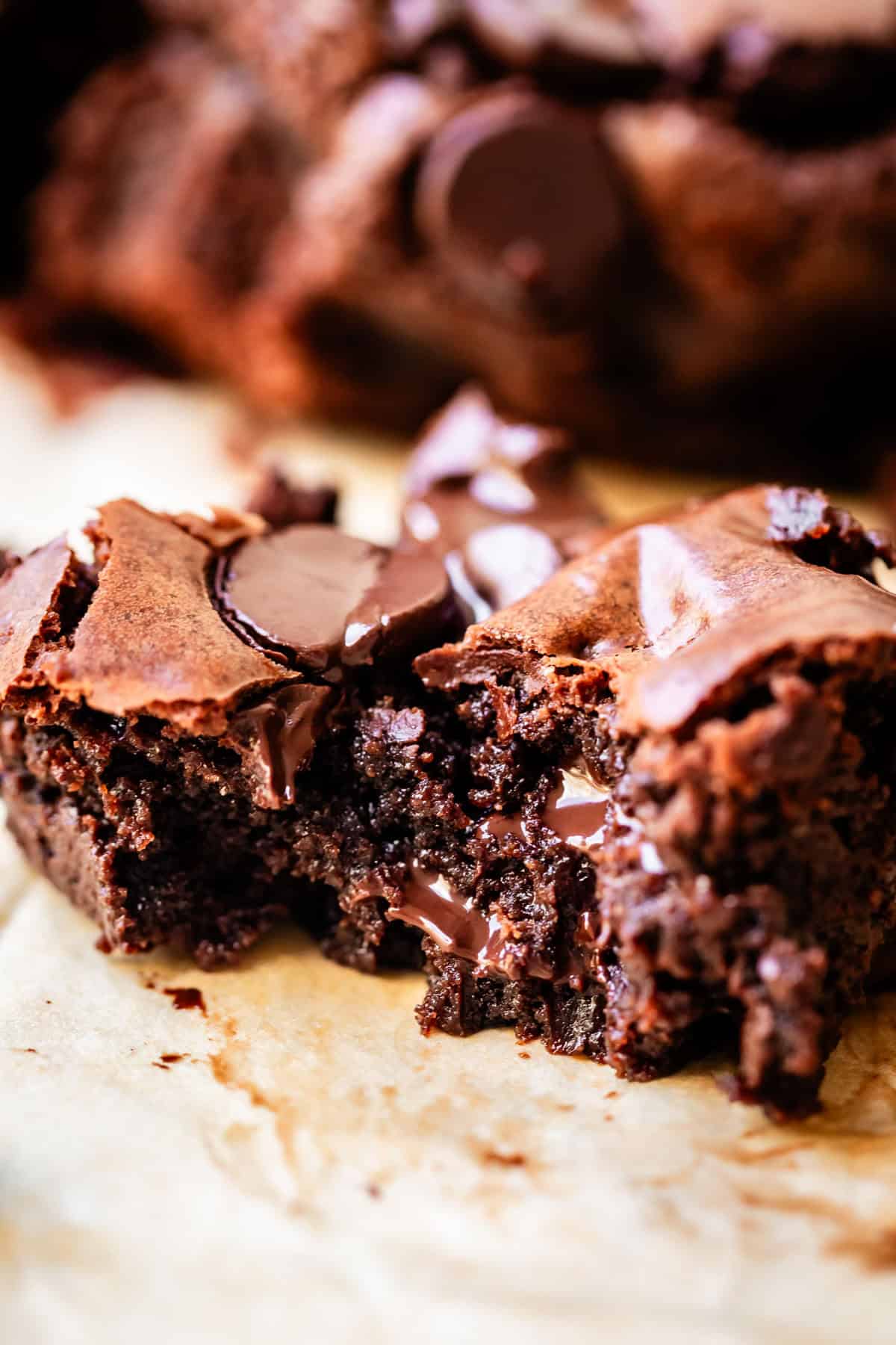 A close-up of a fudgy, chocolate chip gluten free brownie broken in half, showing a gooey, moist center with melted chocolate chunks on top, resting on parchment paper.