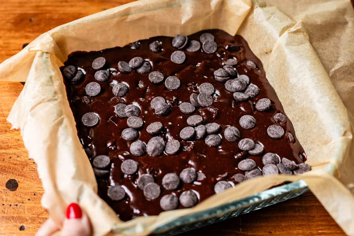 A baking pan lined with parchment paper filled with chocolate brownie batter, topped with chocolate chips, sits on a wooden surface. A hand holds the pan at the corner.