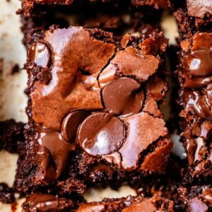 Close-up of a fudgy chocolate brownie topped with melted chocolate chips, showing a cracked, shiny surface and rich, moist texture. Other brownie pieces are partially visible around it.