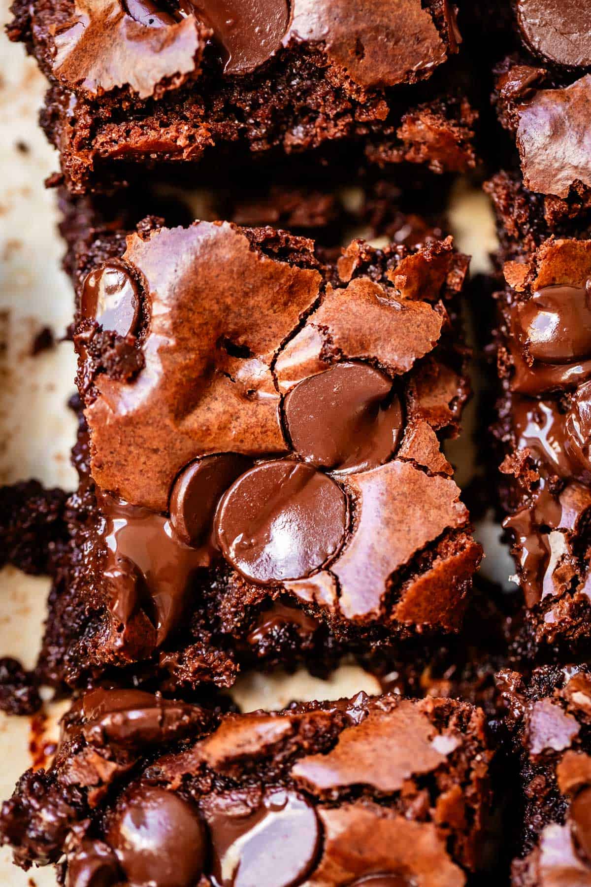 Close-up of a fudgy chocolate brownie topped with melted chocolate chips, showing a cracked, shiny surface and rich, moist texture. Other brownie pieces are partially visible around it.