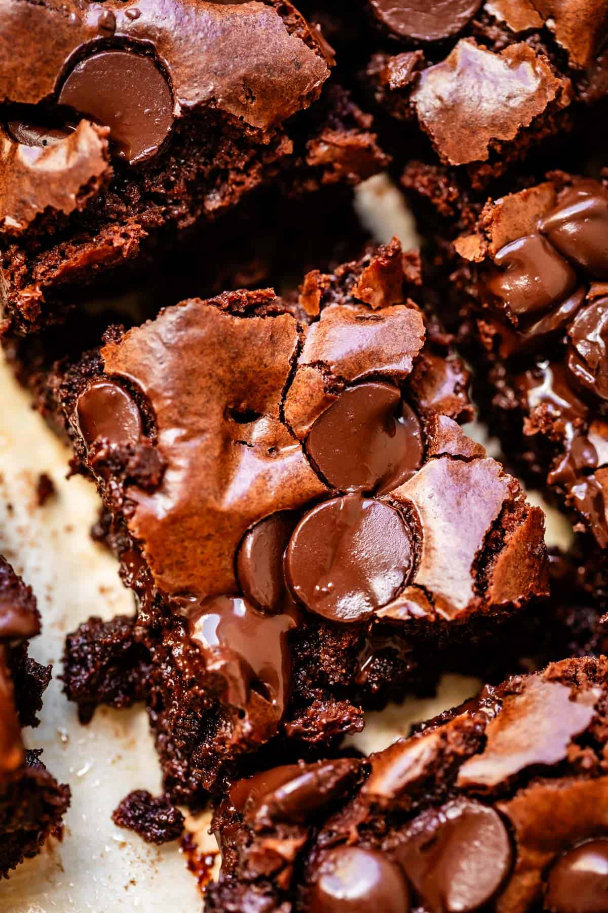Close-up of rich, fudgy brownies topped with melted chocolate chips, showing a cracked, shiny surface and gooey, moist texture inside.