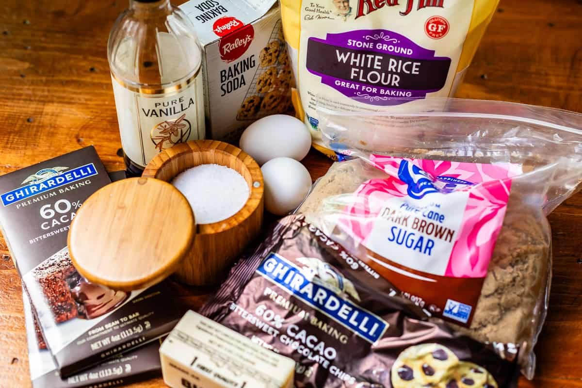 A collection of baking ingredients on a wooden surface, including white rice flour, dark brown sugar, baking soda, pure vanilla, eggs, salt in a wooden container, butter, and two Ghirardelli chocolate bars.