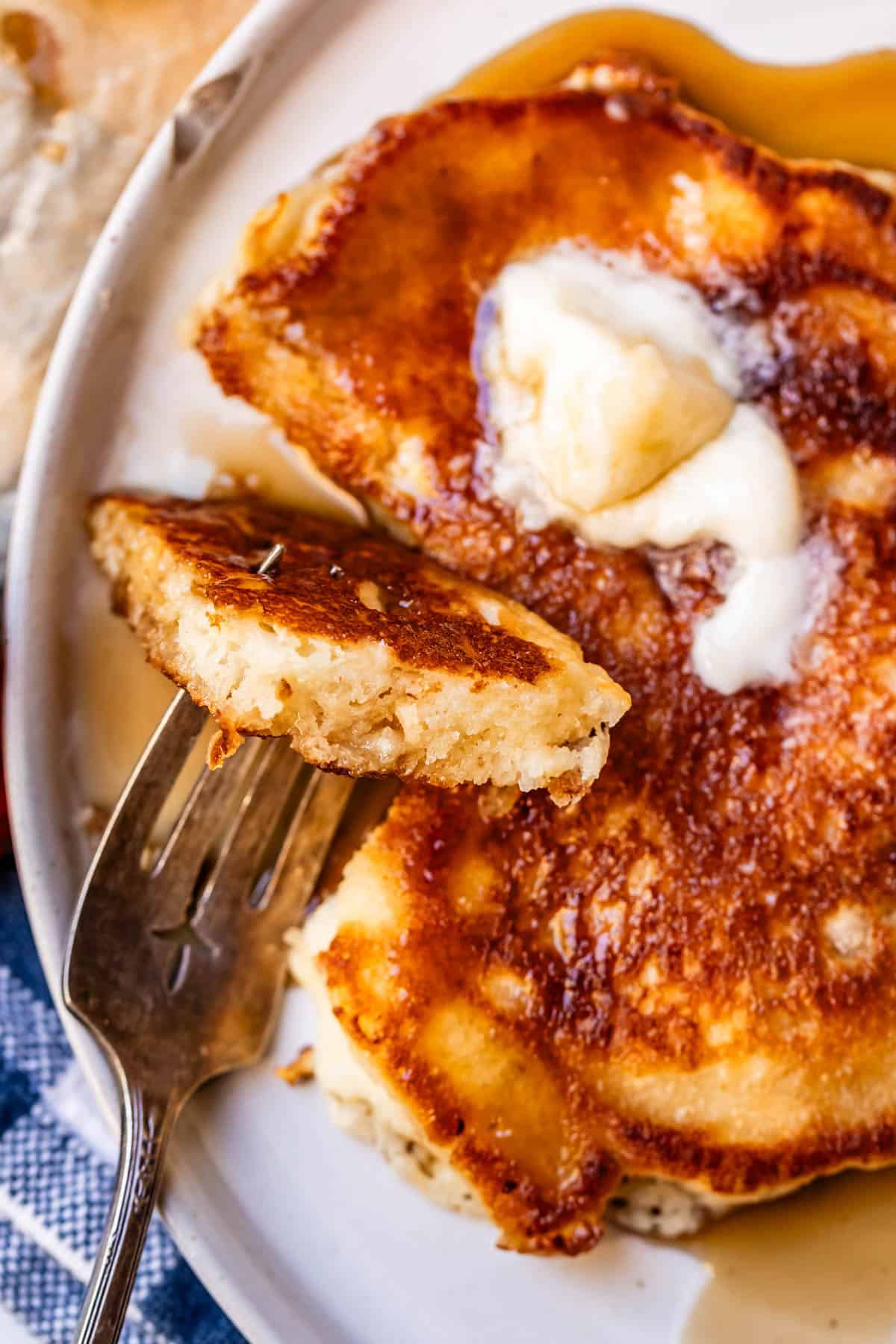 A close-up of a fork holding a bite of fluffy pancake topped with melting butter and syrup on a plate.