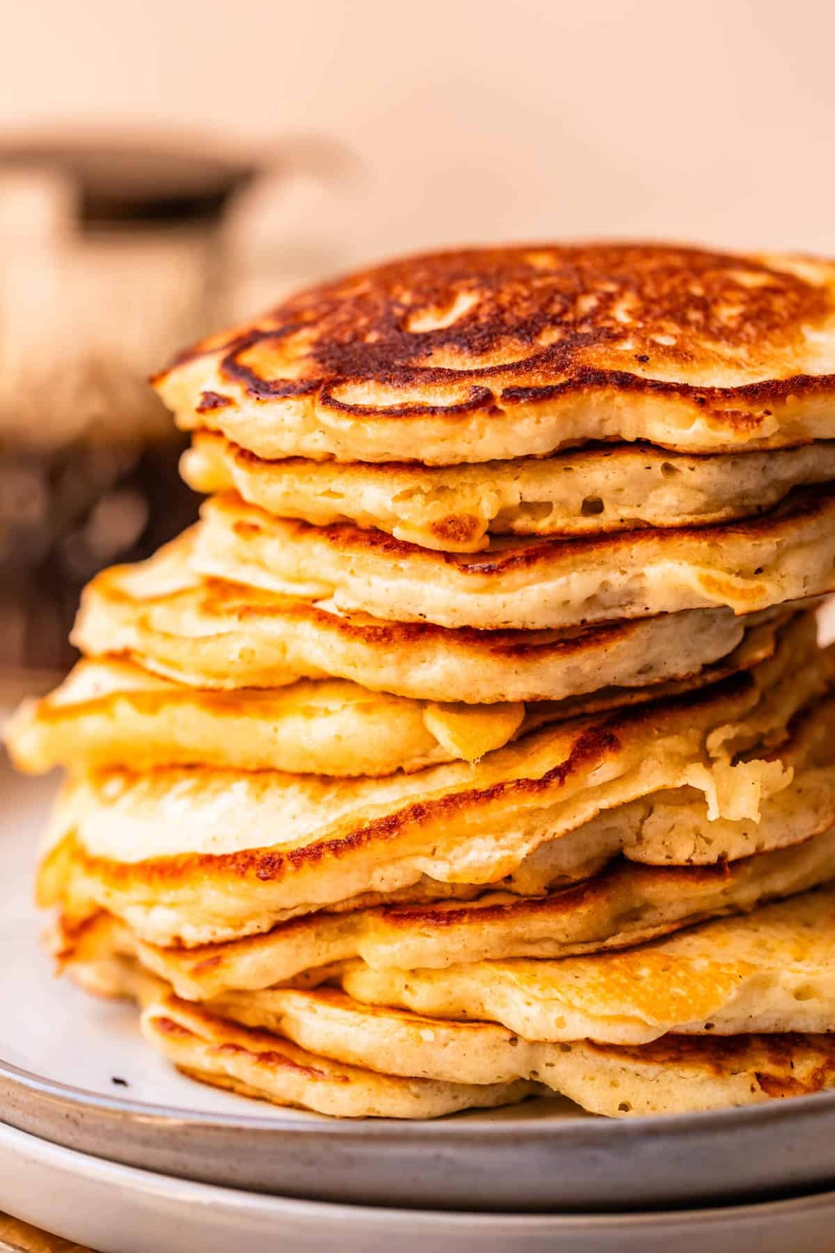 A tall stack of golden-brown, fluffy pancakes sits on a plate, with crisp edges and soft centers visible. The background is softly blurred, highlighting the texture and color of the pancakes.