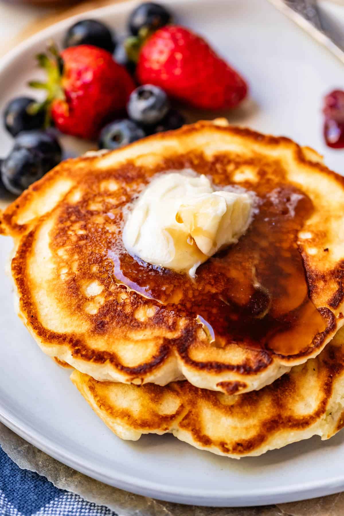 Two golden brown pancakes topped with a melting pat of butter and syrup, served on a white plate with fresh strawberries and blueberries in the background.