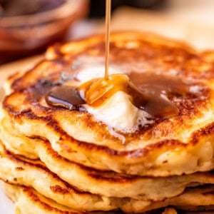 A close-up of a stack of golden-brown pancakes with a pat of melting butter on top, as syrup is being poured over them. The background shows a blurred bowl and dark beverage.