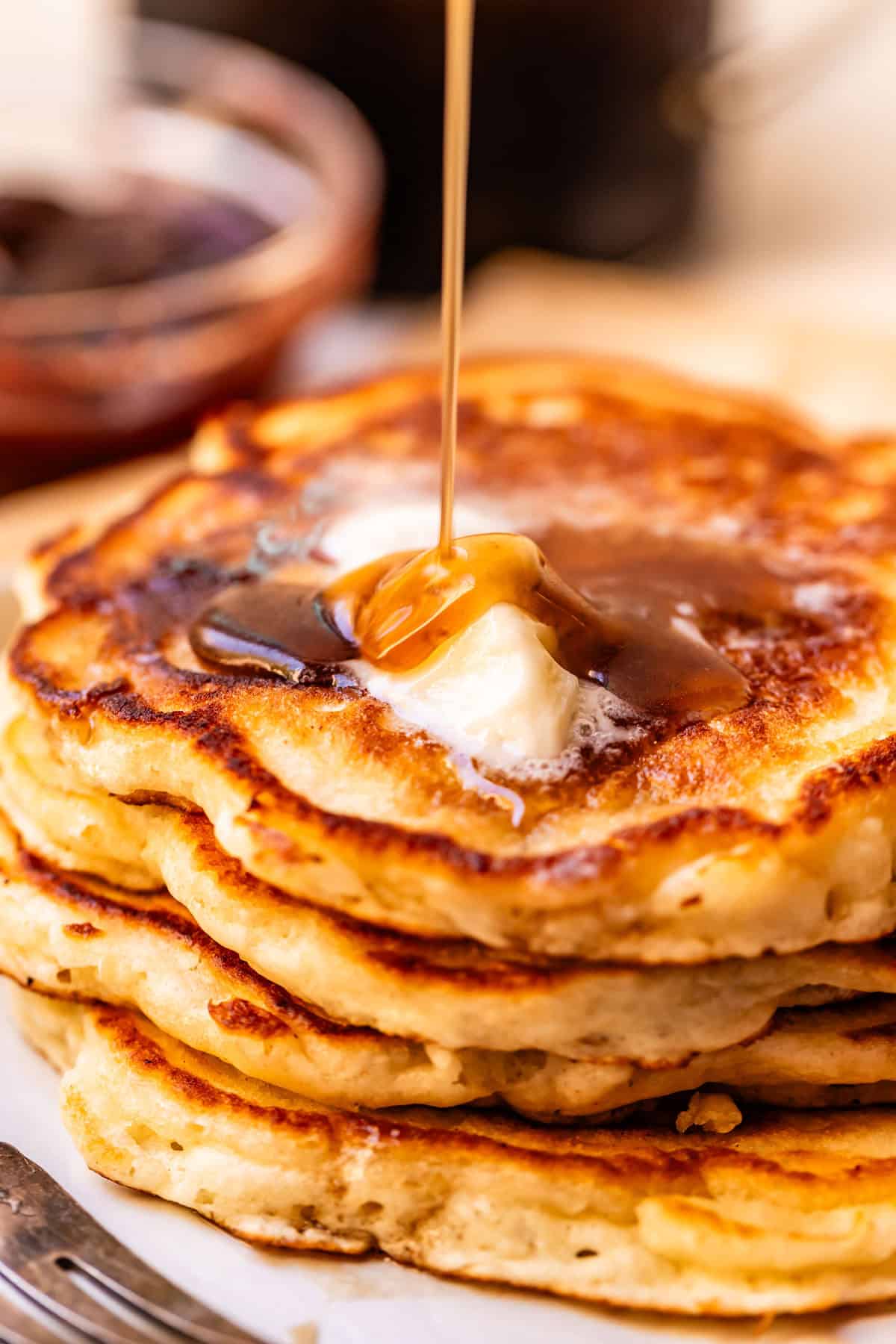 A close-up of a stack of golden-brown pancakes with a pat of melting butter on top, as syrup is being poured over them. The background shows a blurred bowl and dark beverage.
