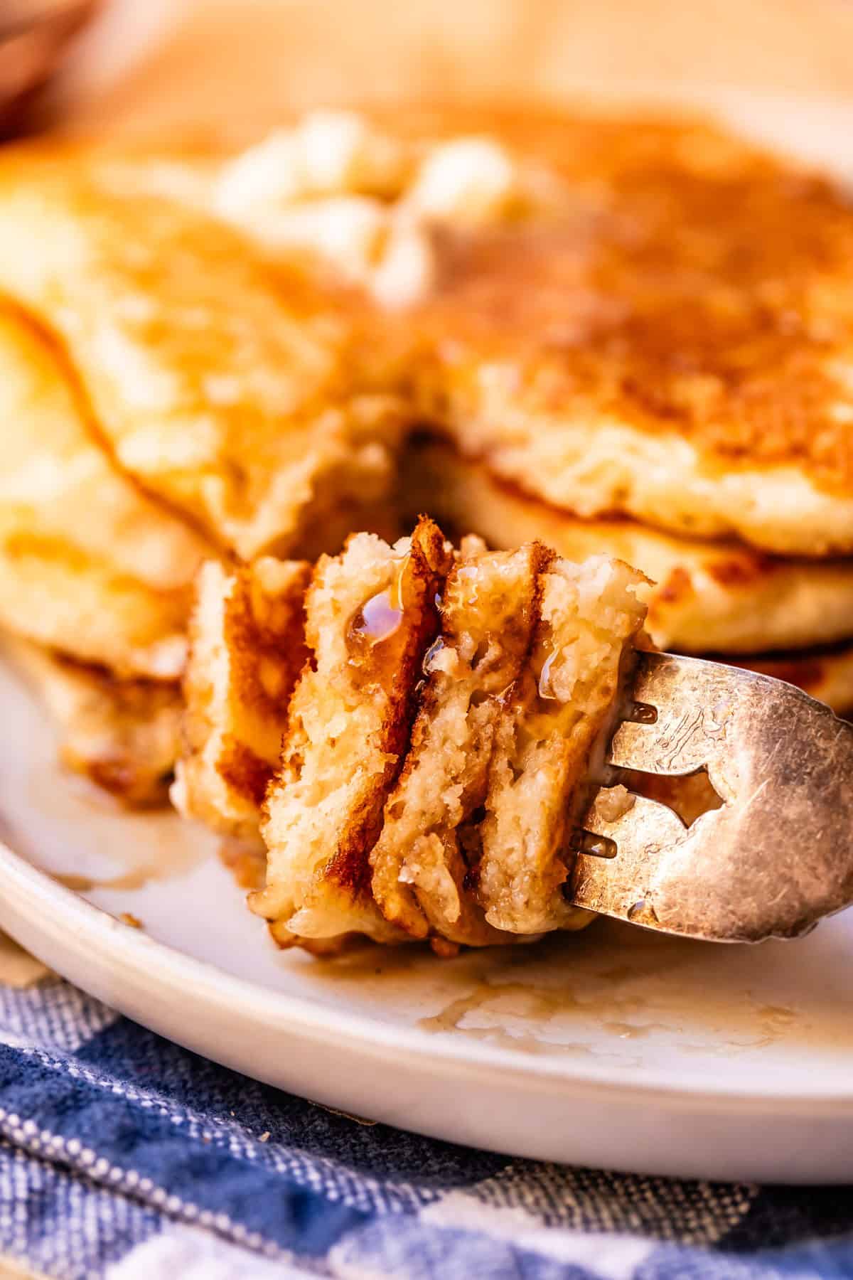 A close-up of a fork holding a bite of fluffy pancakes, topped with syrup and a pat of melting butter, with more pancakes stacked in the background on a plate.