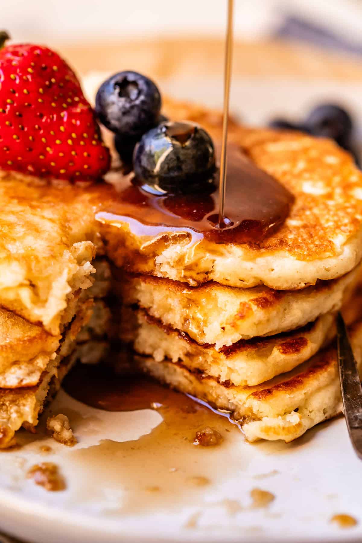 A stack of fluffy pancakes topped with strawberries and blueberries is being drizzled with syrup. One pancake has a bite taken out, revealing the soft, light texture inside.