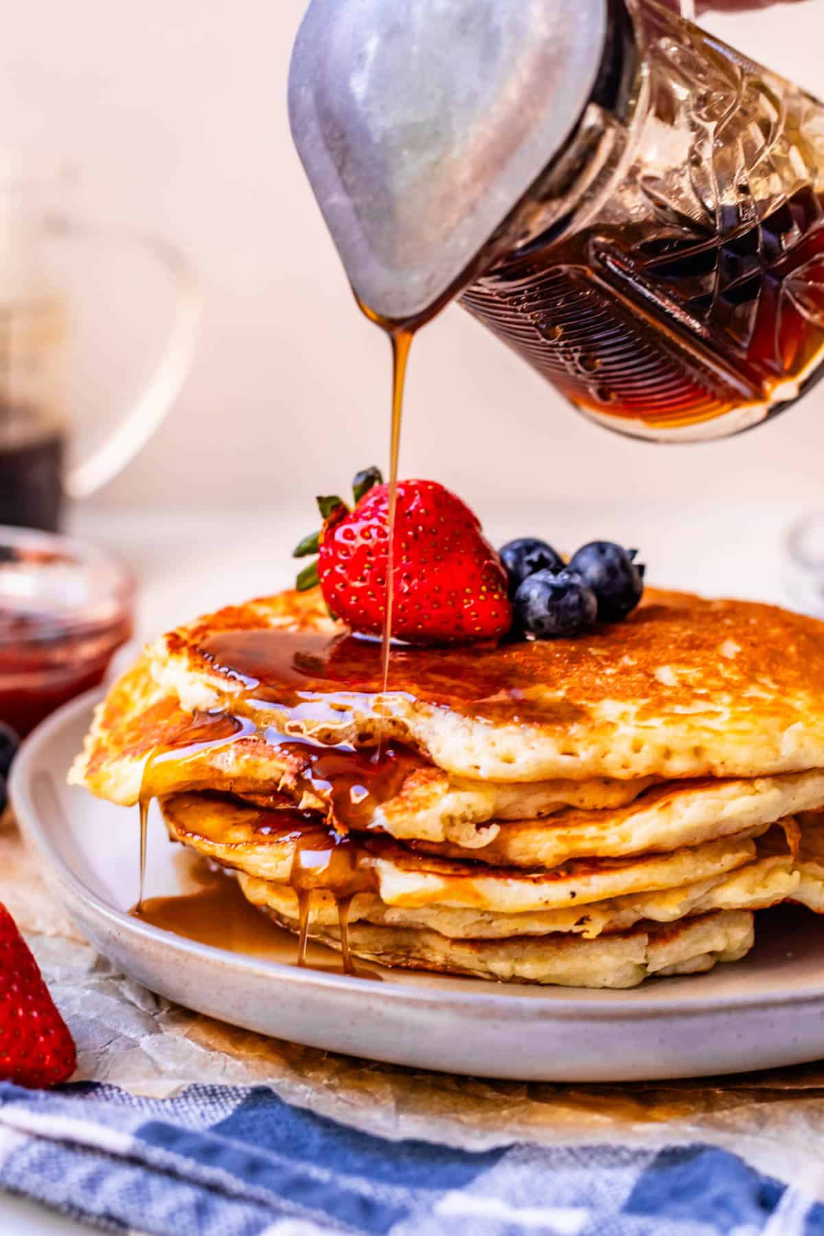 A stack of pancakes topped with strawberries and blueberries is being drizzled with syrup from a pitcher. The pancakes sit on a plate with a blue and white cloth underneath.
