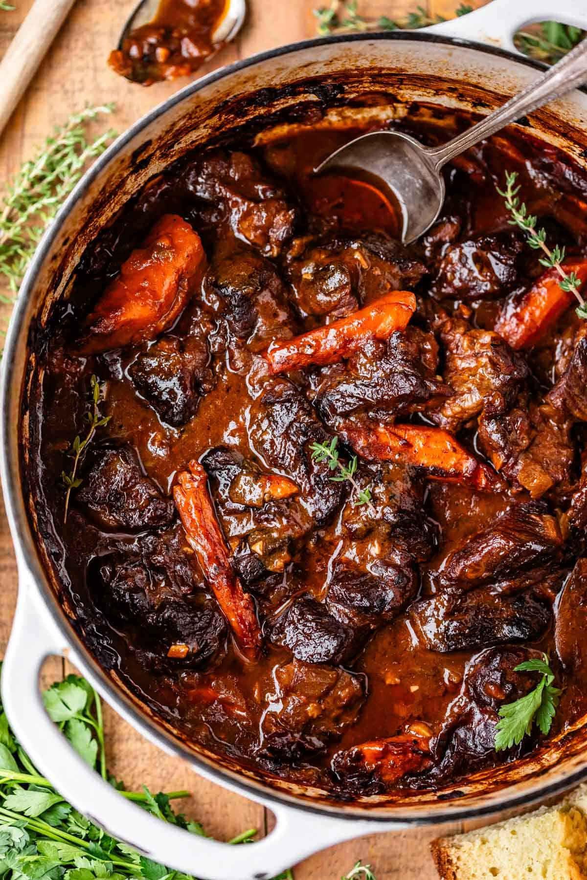 A pot of rich, hearty beef stew with chunks of beef, carrots, and herbs in a thick brown sauce, garnished with fresh parsley, with a spoon resting inside the pot.