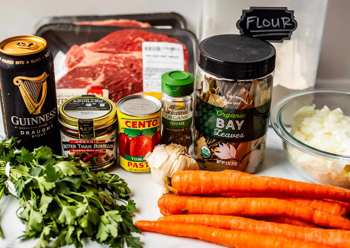 A variety of beef stew ingredients on a counter, including carrots, parsley, garlic, onion, thyme, bay leaves, tomato paste, beef bouillon, flour, a can of Guinness, and a package of beef chuck roast.