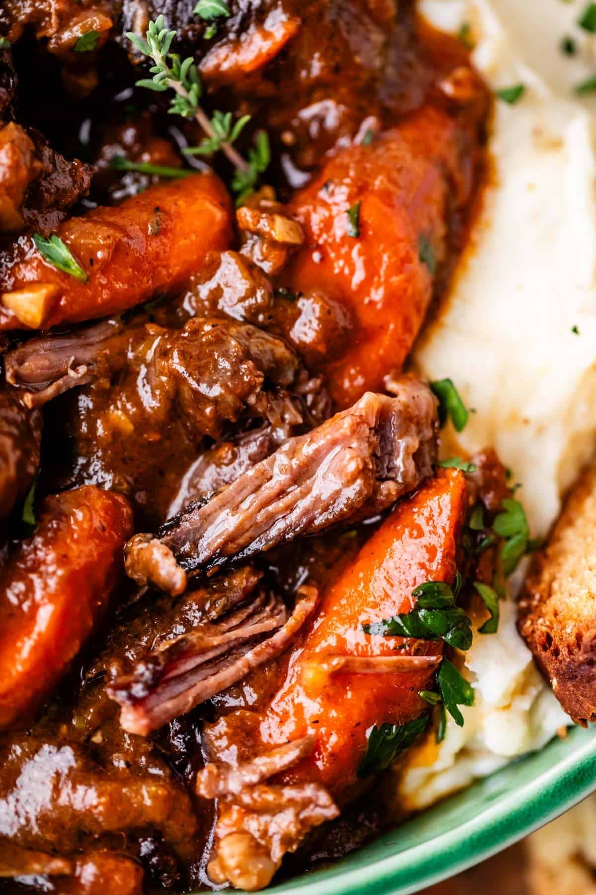 Close-up of tender pot roast with carrots and rich brown gravy, served over creamy mashed potatoes and garnished with fresh herbs. A piece of crusty bread is visible on the side.