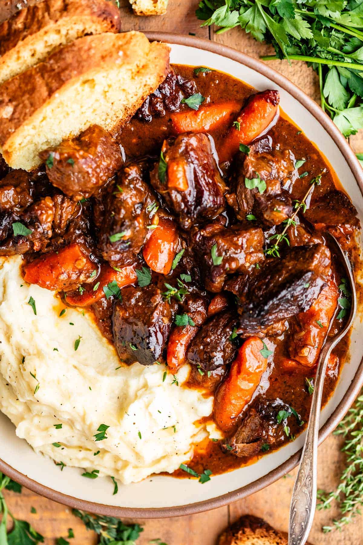 A bowl of beef stew with chunks of beef, carrots, and rich brown gravy is served alongside creamy mashed potatoes, garnished with fresh herbs, and accompanied by slices of bread.