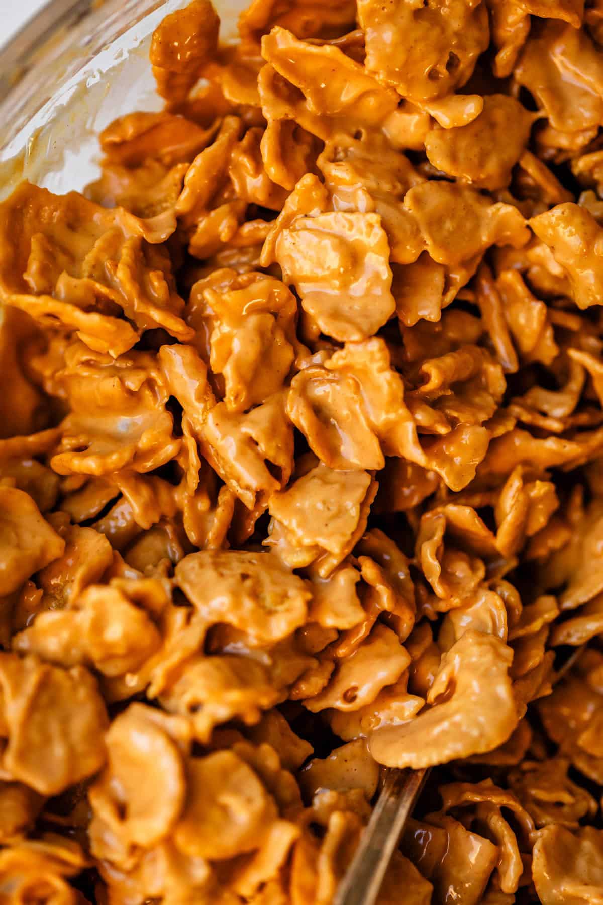 Close-up of cornflakes coated in a glossy, light caramel or peanut butter mixture, creating a crunchy, sweet snack. The flakes are clustered together, highlighting their texture and sticky appearance.