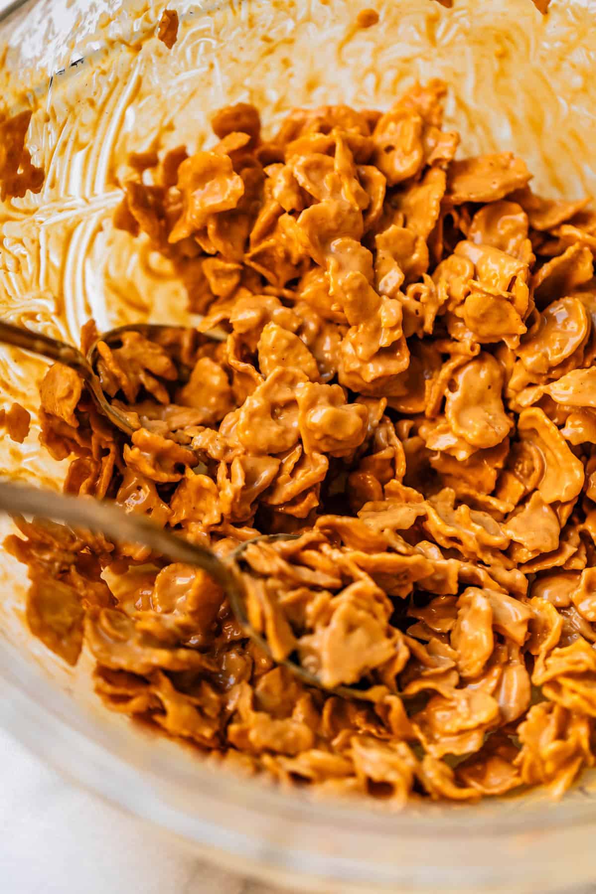 A close-up of cornflakes coated in a creamy, light brown mixture, being stirred with a spoon inside a clear glass bowl.