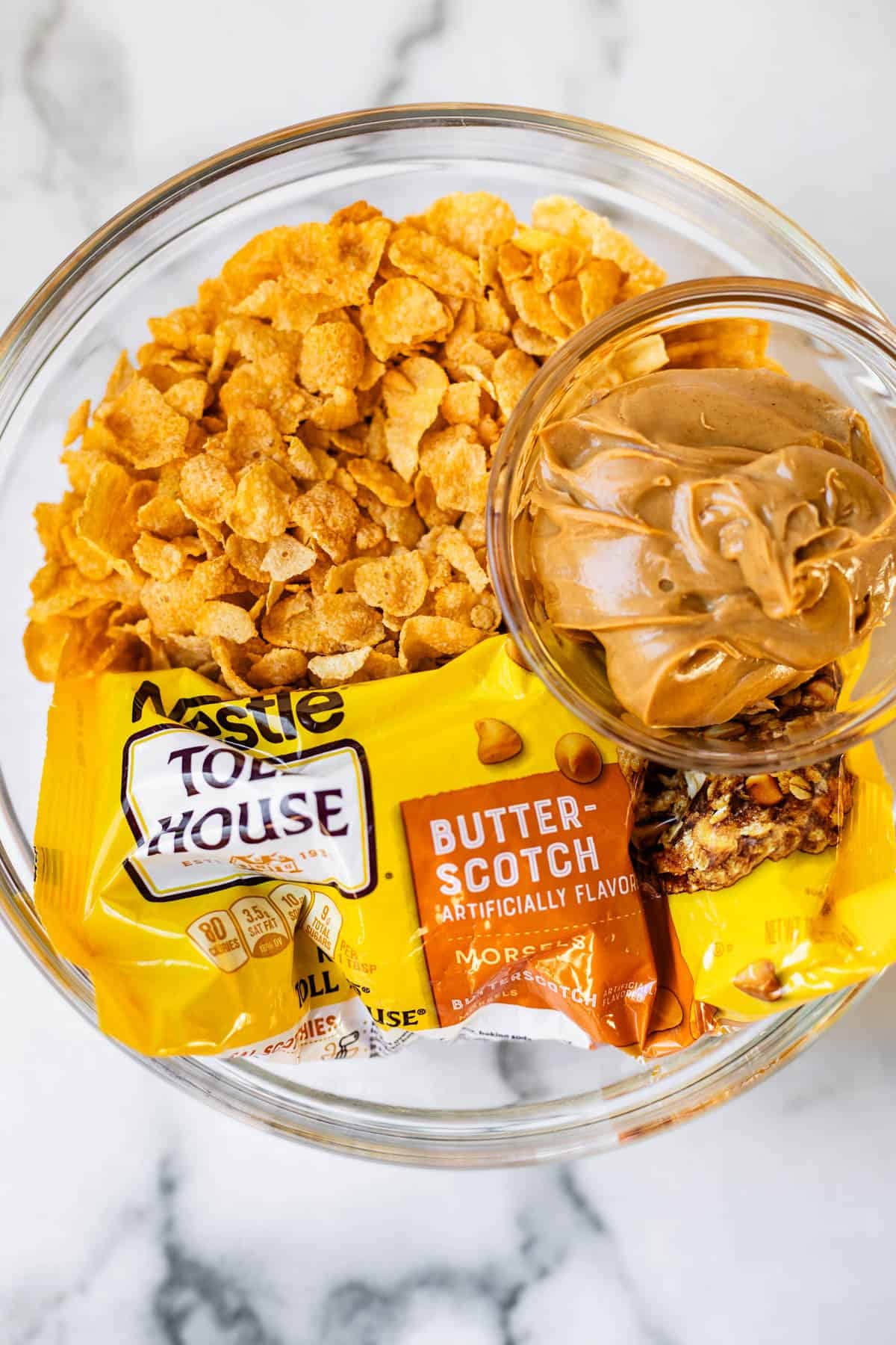 A glass bowl containing cornflakes, a bag of Nestl&eacute; Toll House butterscotch chips, and a small bowl of creamy peanut butter on a white marble surface.
