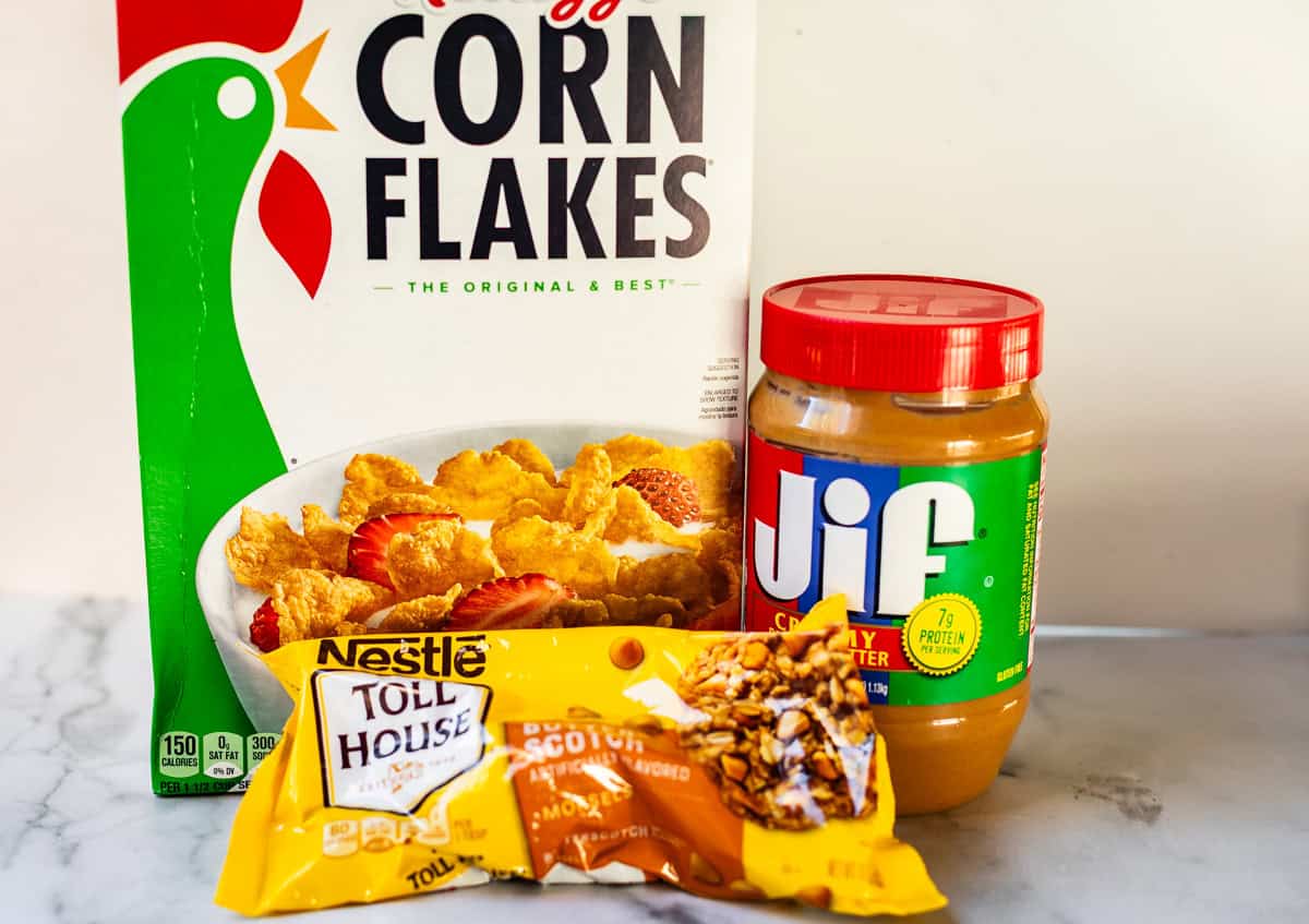 A box of Kelloggs Corn Flakes, a jar of Jif creamy peanut butter, and a bag of Nestl&eacute; Toll House butterscotch morsels on a marble surface.