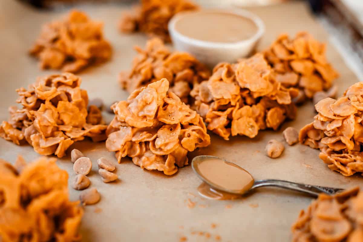 Clusters of cornflakes coated in a light brown peanut butter mixture are arranged on parchment paper, with a spoon of peanut butter and a small bowl in the background.