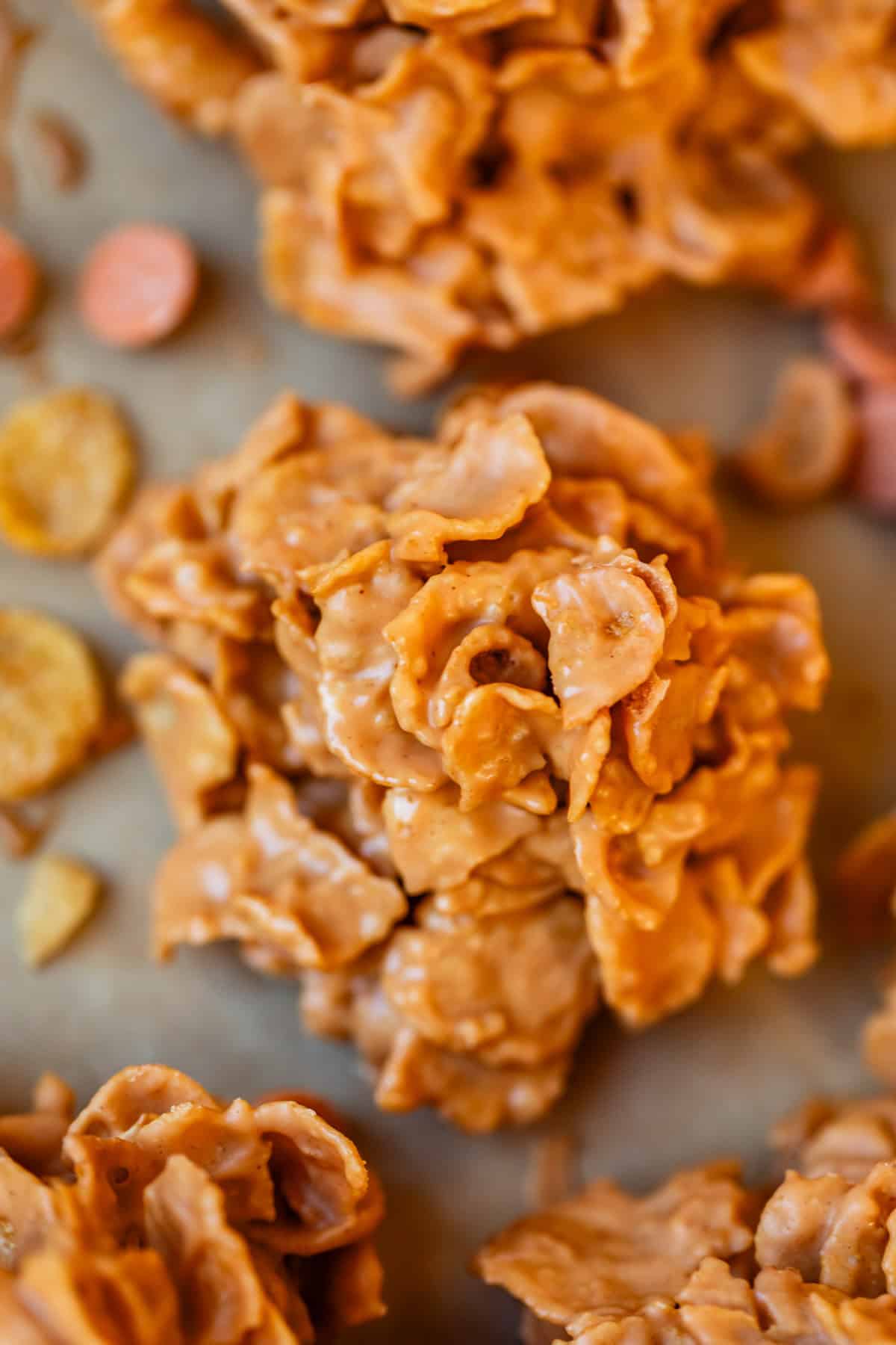 Close-up of a no-bake butterscotch haystack cookie made with cornflakes, coated in a creamy, light brown butterscotch mixture. Other pieces and scattered cornflakes are visible in the background.