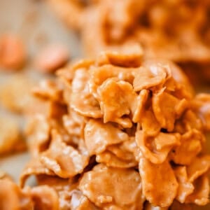 Close-up of a cluster of golden, crispy cornflakes coated in a creamy, caramel-colored mixture, likely a no-bake treat or dessert snack, with more clusters blurred in the background.