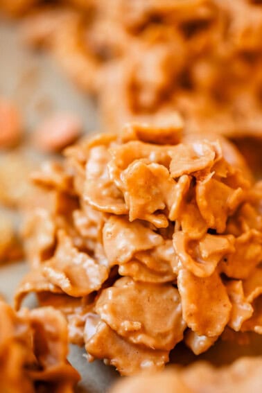 Close-up of a cluster of golden, crispy cornflakes coated in a creamy, caramel-colored mixture, likely a no-bake treat or dessert snack, with more clusters blurred in the background.