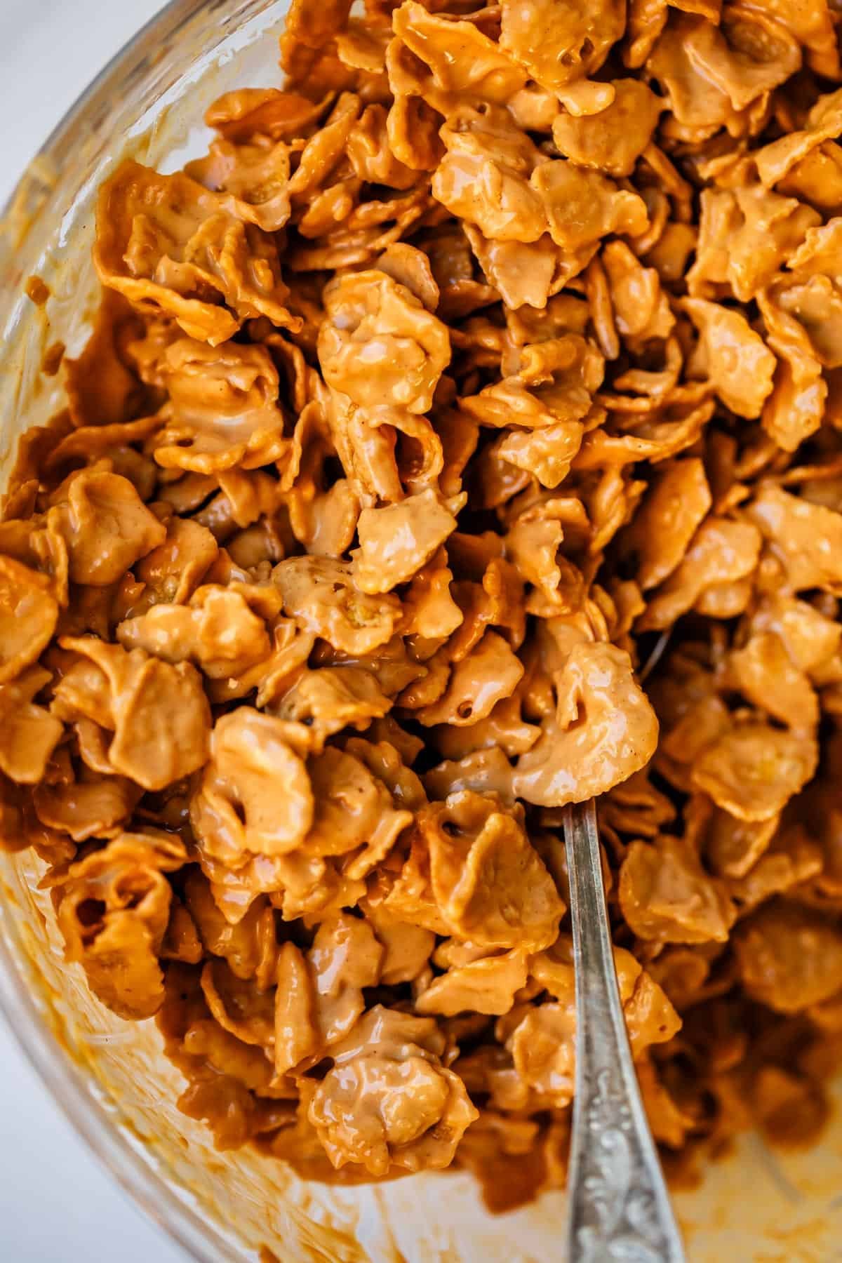 Close-up of a bowl filled with cornflakes coated in a glossy, caramel-colored mixture, with a metal spoon partially visible on the right side.