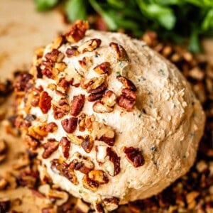 A round cheese ball coated with chopped pecans sits on a wooden surface, with fresh green herbs in the background.