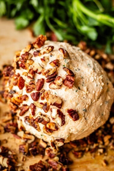 A round cheese ball coated with chopped pecans sits on a wooden surface, with fresh green herbs in the background.