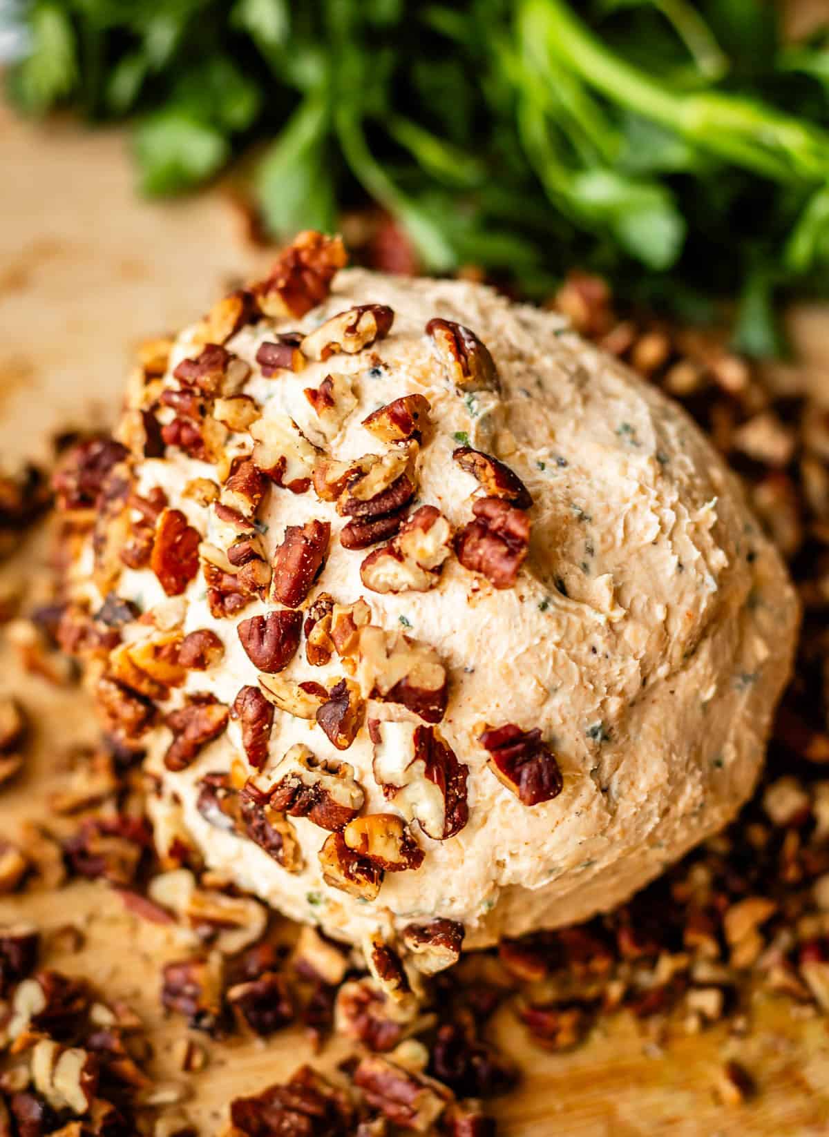 A round cheese ball coated with chopped pecans sits on a wooden surface, with fresh green herbs in the background.