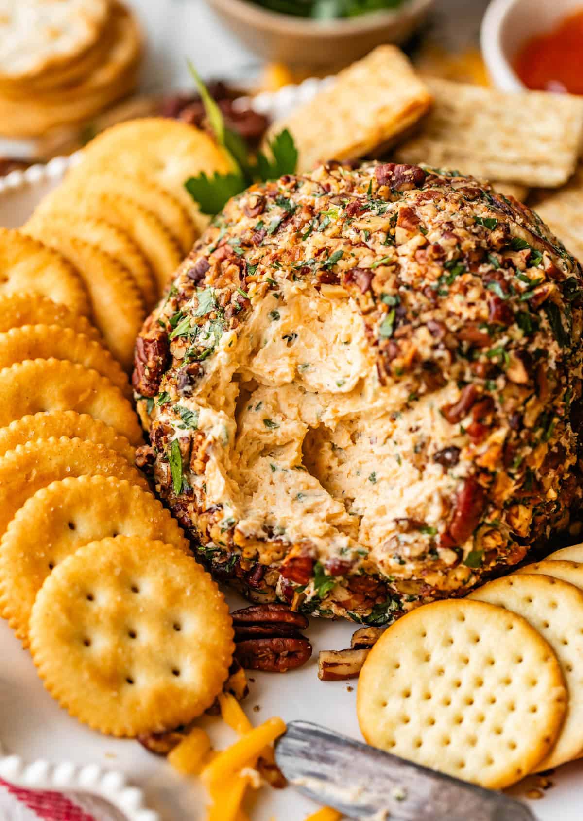 A close-up of a cheese ball coated in chopped parsley and pecans, surrounded by round crackers on a white platter, with some cheese spread missing from the center.