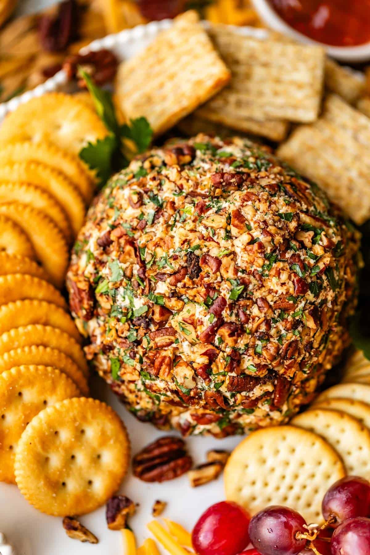 A round three cheese cheese ball coated with chopped pecans and parsley is surrounded by an assortment of crackers, grapes, and fresh parsley on a white platter.