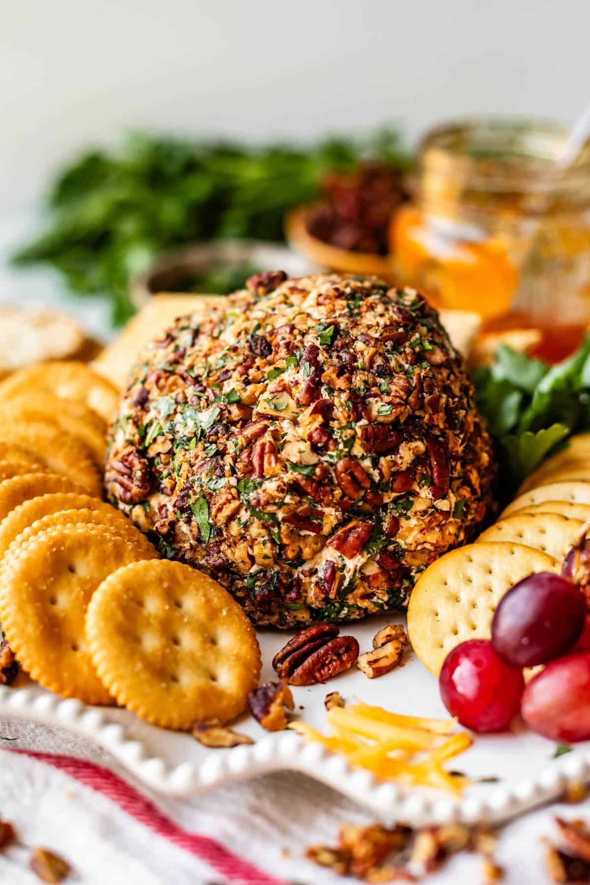 A pecan-crusted cheese ball surrounded by round crackers, grapes, shredded cheese, and fresh parsley on a white platter, with a jar of apricot jam in the background.