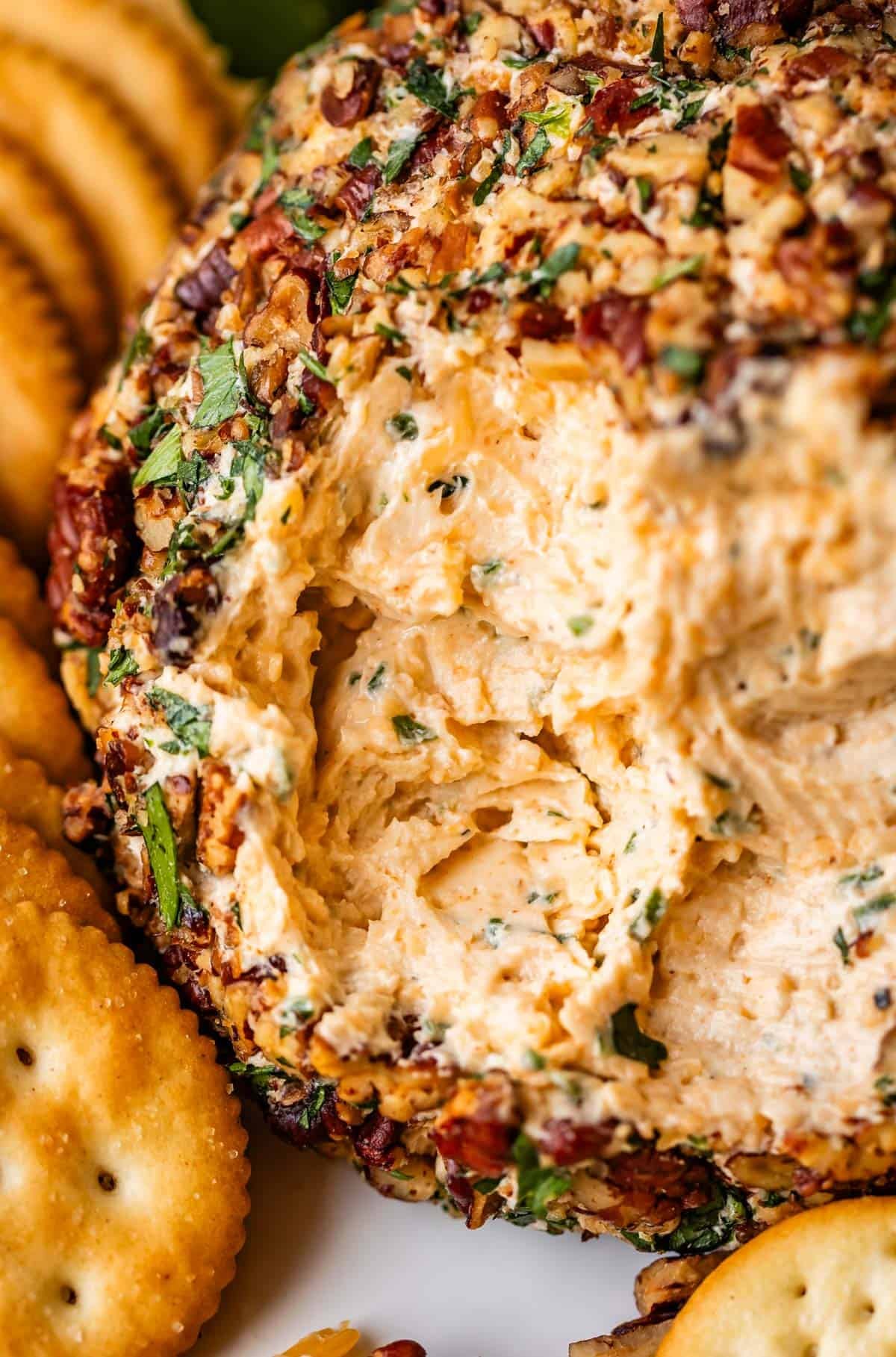 A close-up of a classic cheese ball coated in chopped pecans and herbs, with a portion scooped out, surrounded by round crackers on a white plate.