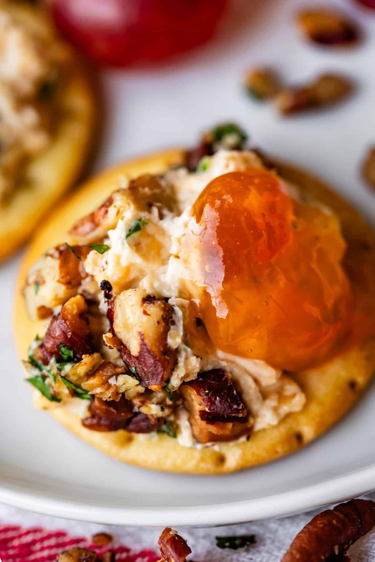 A close-up of a cracker topped with cheese ball spread, chopped nuts, parsley, and a dollop of apricot jam, on a white plate with scattered pecan pieces nearby.