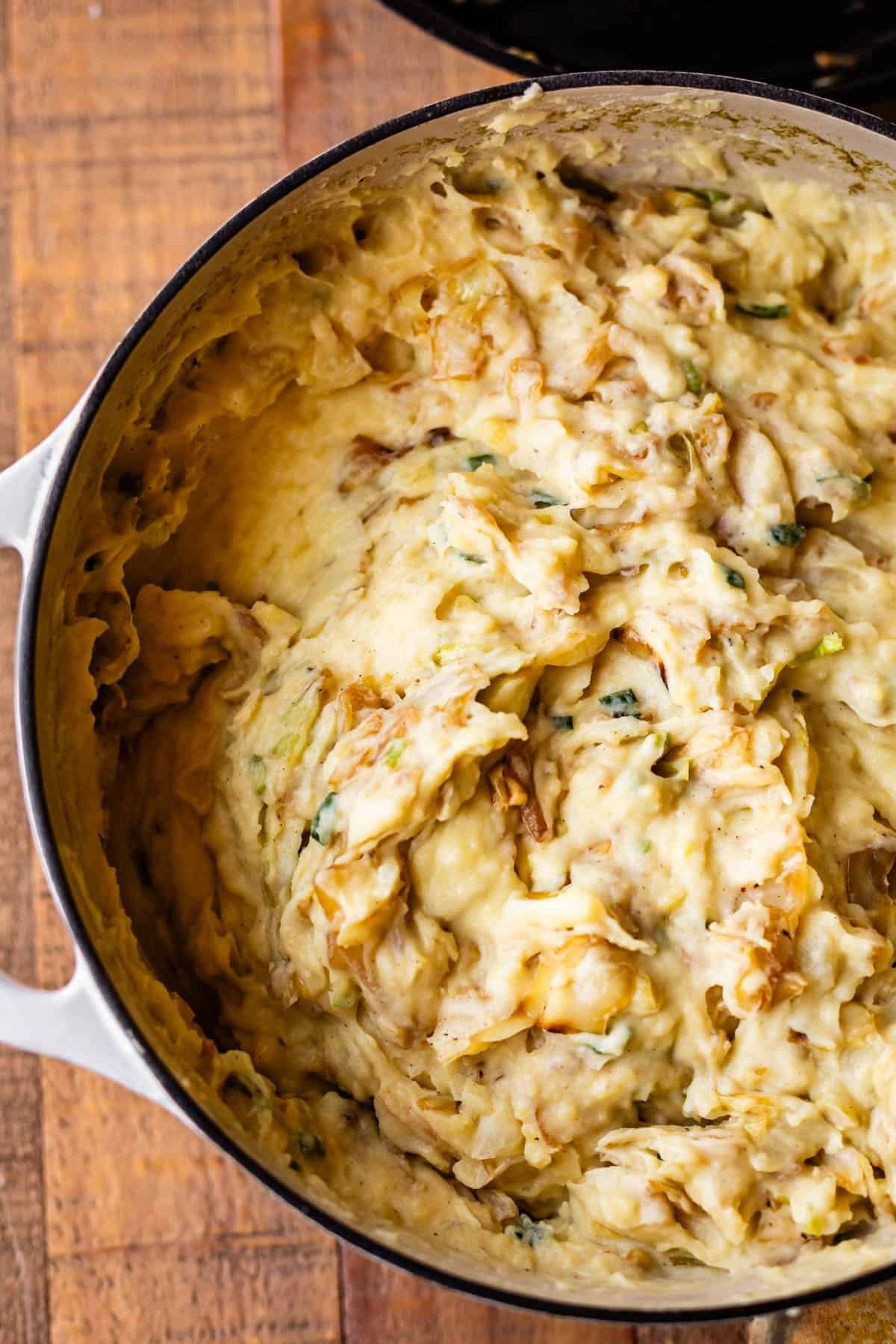A pot filled with creamy mashed potatoes mixed with caramelized onions and herbs, sitting on a wooden surface.