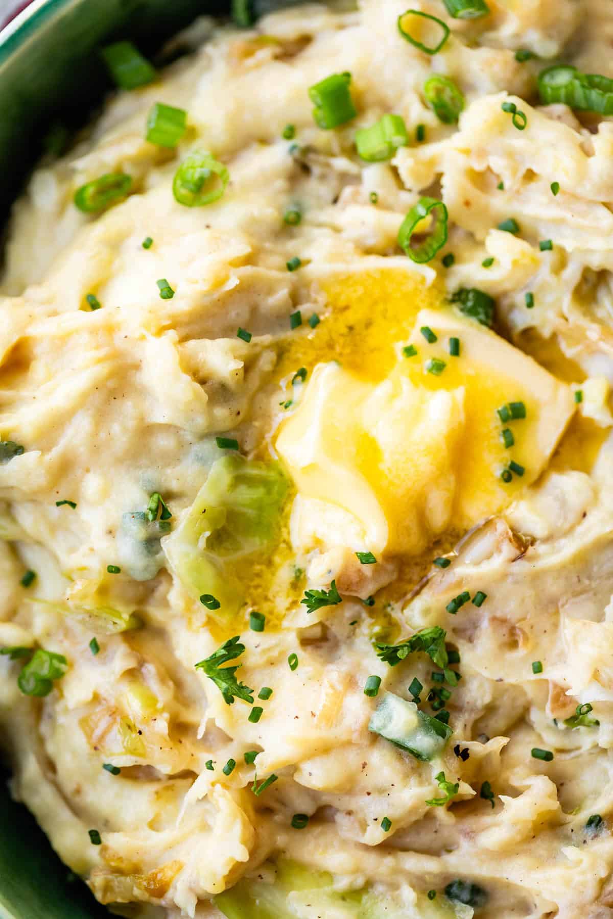 Close-up of creamy mashed potatoes with green onions, topped with a pat of melted butter and sprinkled with chopped chives.