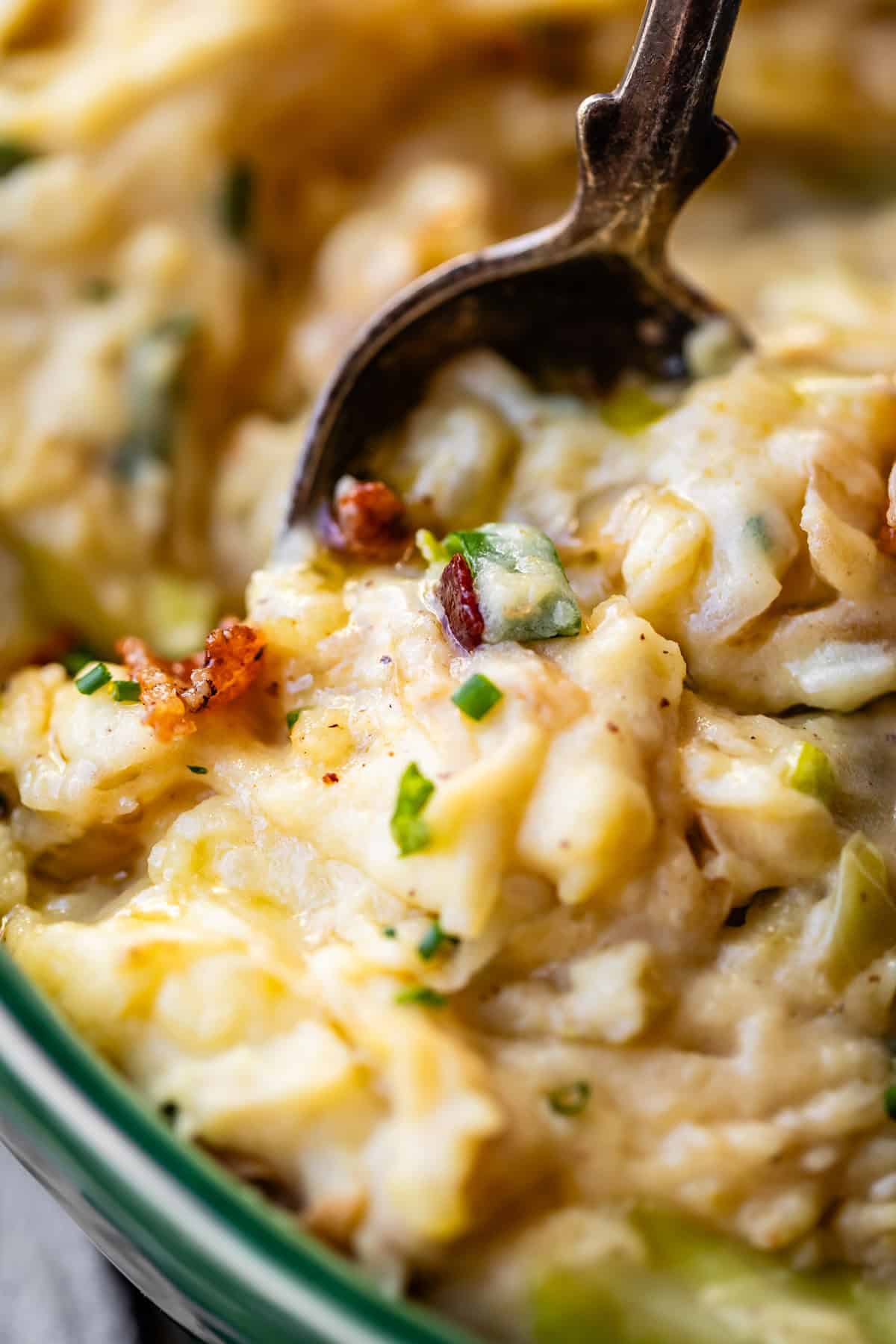 Close-up of creamy mashed potatoes mixed with chopped chives and crispy bacon bits, being scooped with a metal spoon. The texture appears rich and slightly chunky.