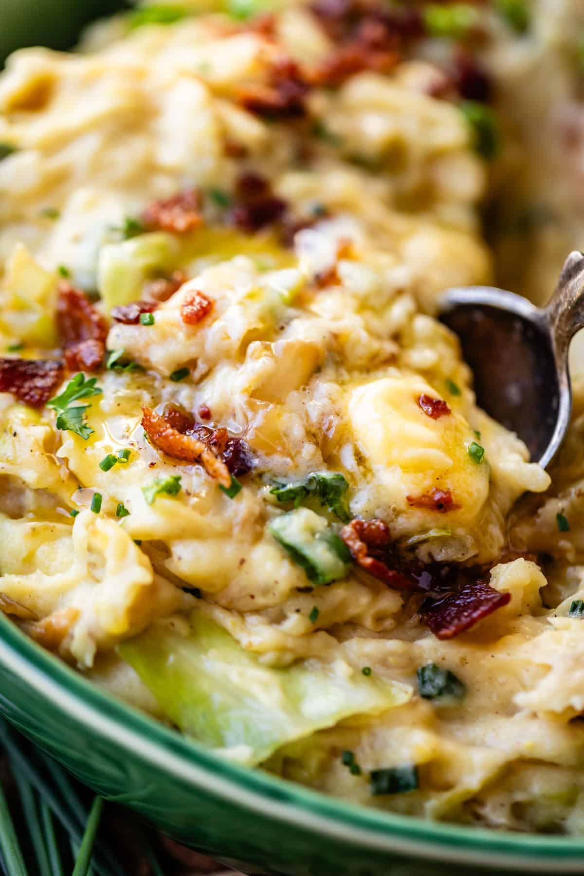 Close-up of a creamy casserole topped with melted cheese, crispy bacon bits, and chopped herbs, with a spoon scooping out a portion from a green dish.