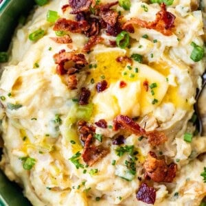 A bowl of creamy mashed potatoes mixed with green onions and topped with crispy bacon bits, melted butter, and chopped chives.