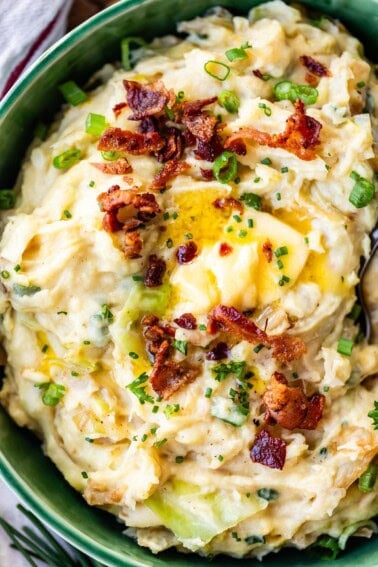 A bowl of creamy mashed potatoes mixed with green onions and topped with crispy bacon bits, melted butter, and chopped chives.
